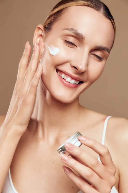 Smiling woman applying face cream on cheek, close up portrait, natural skin care routine, soft lighting, relaxed expression, smooth skin, neutral background.