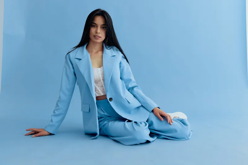 young woman sitting on floor in blue pantsuit against seamless blue background, long dark hair, casual pose, modern fashion outfit