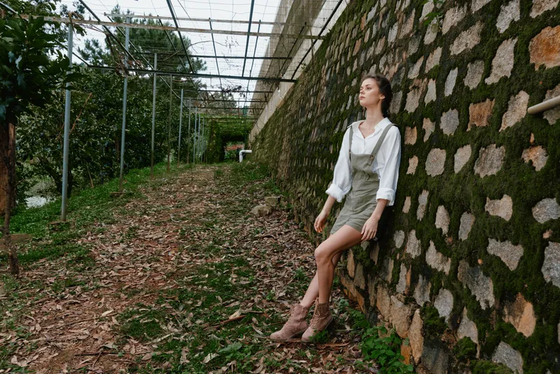 Beautiful young woman in casual outfit enjoying nature, surrounded by greenery and textured stone wall in a peaceful outdoor setting