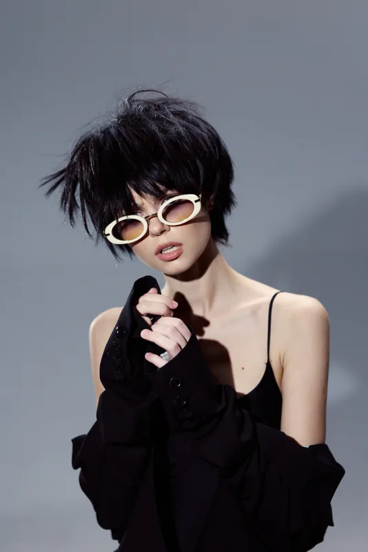 Stylish woman with short black hair and white sunglasses wearing a black outfit posing with hands clasped, isolated on gray background.