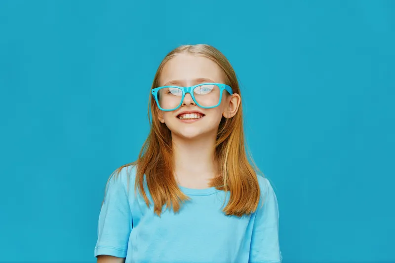 Cheerful young girl with blonde hair wearing blue glasses and a light blue shirt against a vibrant turquoise background, radiating positivity and joy