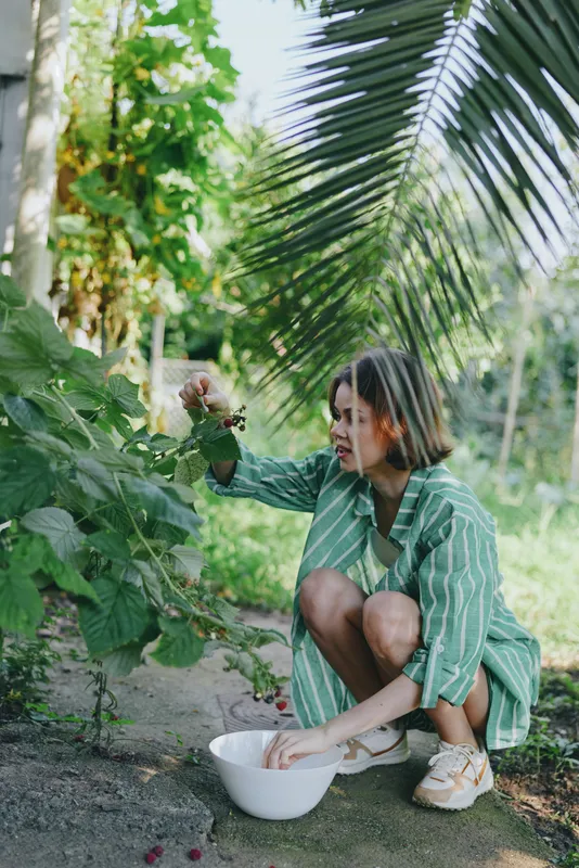 Woman, garden, planting, a cheerful woman kneels beside lush plants, hands near a bowl, gently tending foliage under natural light, a calm outdoor scene full of greenery and care