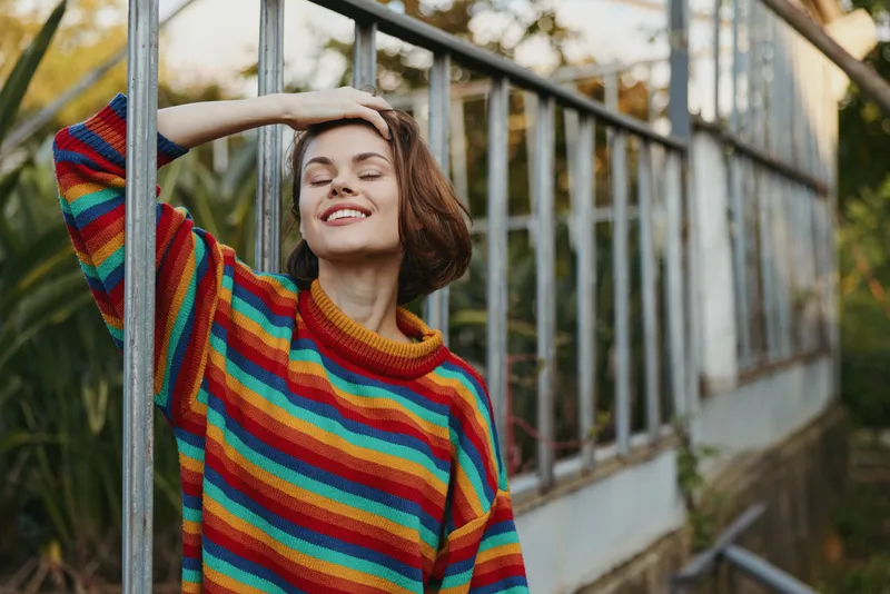 woman striped sweater smiling greenhouse leaning outdoor autumn young adult: joyful 25-year-old female with short brown hair in colorful knit sweater, relaxed pose by vintage greenhouse in golden