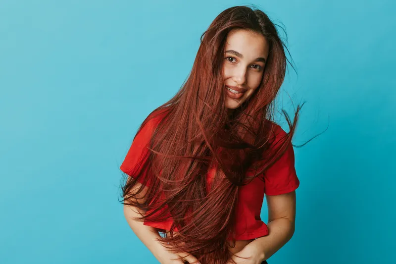 Young woman with long brown hair wearing red shirt smiling and posing against blue background. Casual portrait with natural expression and vibrant color contrast in studio setting.