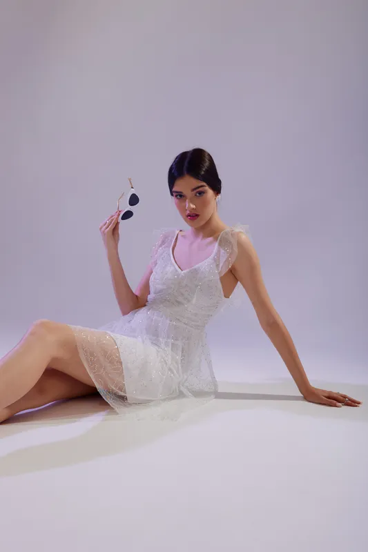 Young woman in a white lace dress poses sitting on the floor holding sunglasses. Studio shot with soft lighting on a light background. Concept of beauty, fashion, and elegance.