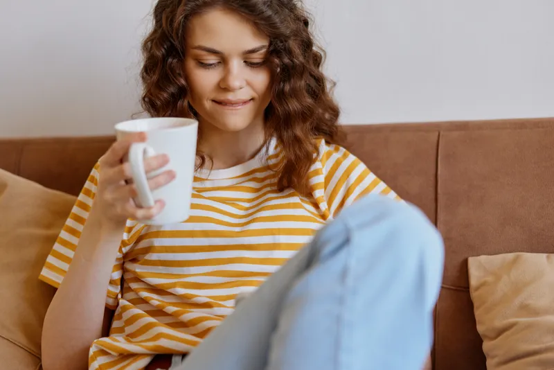 A relaxed woman sits on a sofa at home, sipping from a mug in a striped tee and jeans. A cozy lifestyle scene highlighting comfort, routine, and self care.
