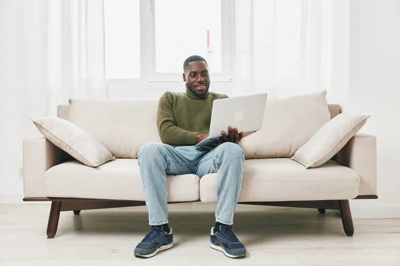 Smiling young Black man working on a laptop in a cozy living room, wearing casual clothing, with soft lighting and a neutral color palette