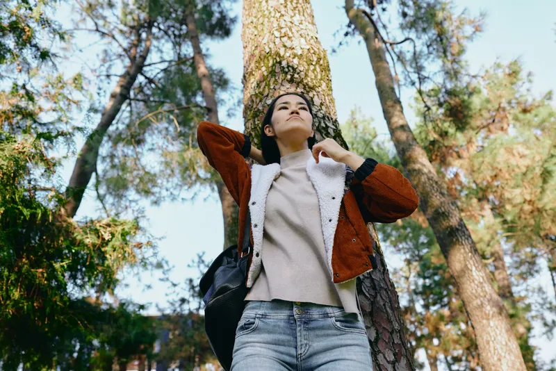Woman, forest, outdoors, jacket, sweater, backpack, casual, nature, lifestyle, autumn, tall trees frame the scene, sky peeks through leaves, warm sunshine, tranquil mood, exploration and renewal in