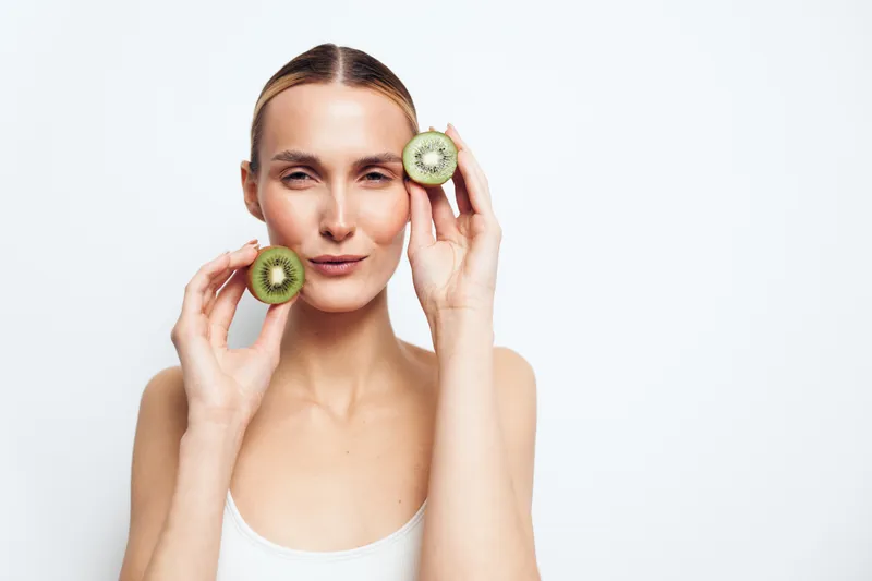Young woman holding two halves of fresh kiwi fruit near face smiling gently clean skin white background minimal style natural beauty concept