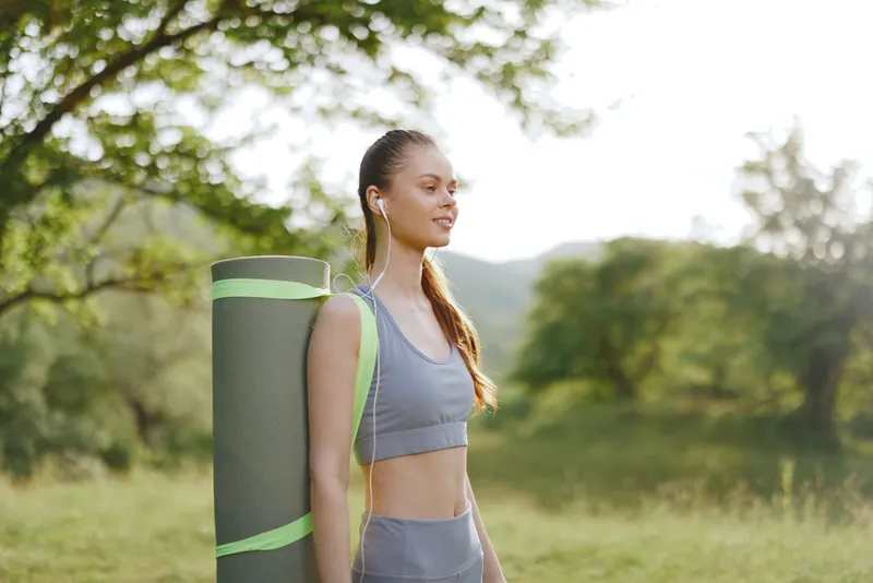 Young woman in sportswear carrying a yoga mat, smiling and enjoying a sunny day in nature, embodying a healthy lifestyle and fitness concept