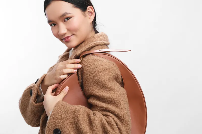 Asian woman wearing a brown coat carries a round leather backpack in a clean studio setting. She smiles softly, creating a warm, stylish everyday travel vibe and approachable professional aura.