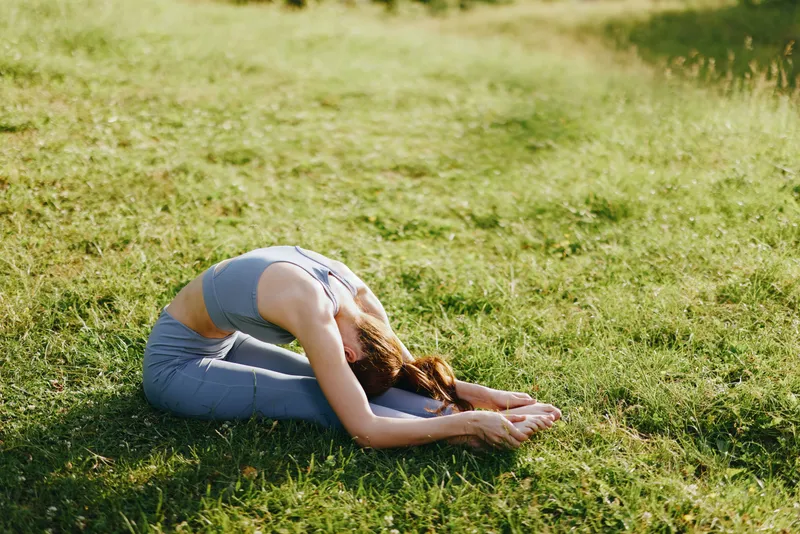 Young woman practicing yoga outdoors, demonstrating flexibility in a serene green field, wearing a comfortable blue outfit, expressing calmness and focus during her routine