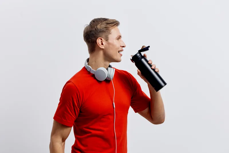 Smiling young man in red t-shirt with headphones holding black water bottle against plain light gray background studio portrait. People lifestyle concept