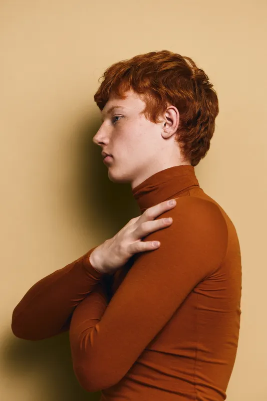 Profile of young redhead man wearing brown turtleneck posing with crossed arms against beige background in studio for fashion portrait.