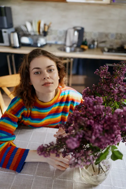 Woman lifestyle home scene with a relaxed mood, seated at a kitchen table, bouquet of lilacs nearby, wearing a colorful striped sweater, natural light highlighting calm daily moments