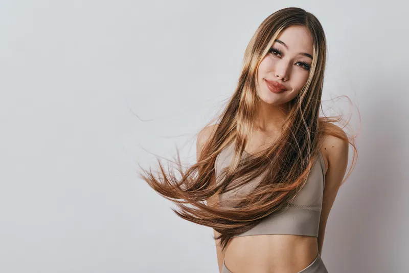 Young woman with long flowing hair smiling at camera wearing a crop top on light background with natural makeup and confident pose in studio.