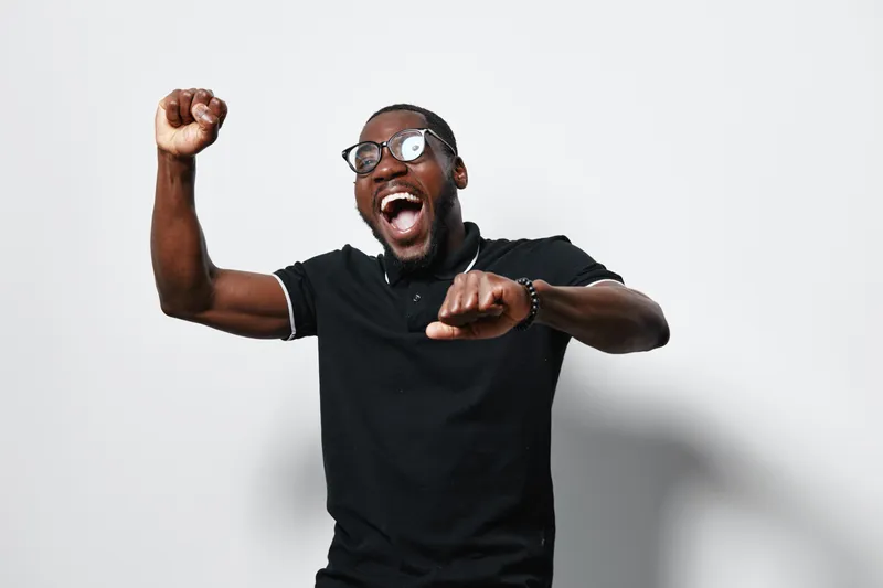 African American man with dark skin wearing glasses and black shirt celebrating with raised fists and joyful expression on white background. Happiness and excitement concept.