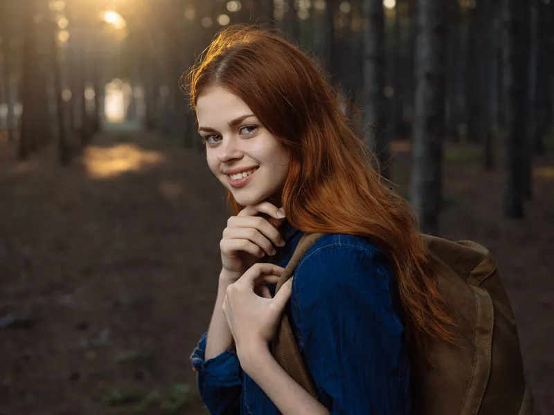 Женщина в синей рубашке с рюкзаком за спиной, вид сбоку, в сосновом лесу
