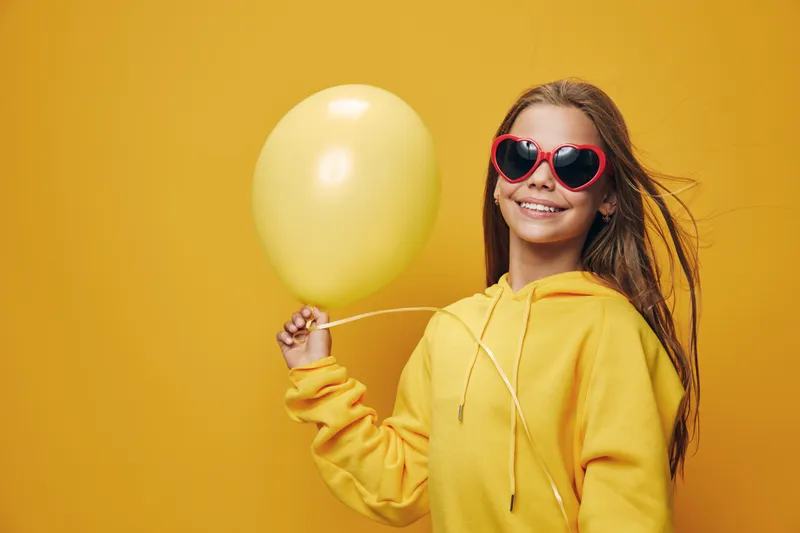 Girl wearing yellow hoodie and heart shaped sunglasses holding yellow balloon smiling on yellow background with flowing hair for childhood and happiness concept.