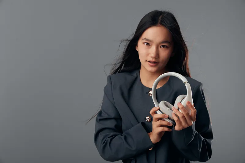 Young woman holding white headphones with both hands against grey background for music and technology concept. Female with long dark hair wearing dark jacket looking at camera.