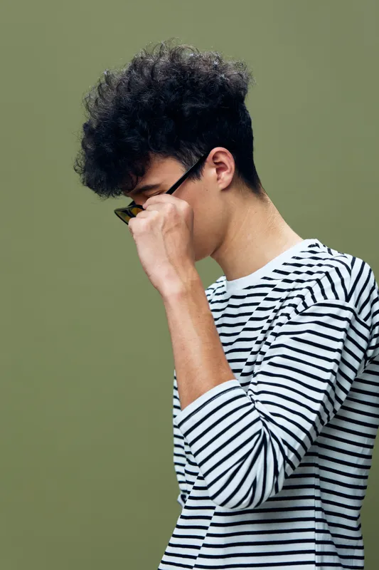 young man adjusting glasses wearing striped shirt with curly hair on olive background, casual style, thoughtful mood, studio portrait, clean composition