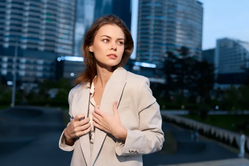 Confident young woman in beige blazer posing in modern urban environment with skyscrapers at dusk, business concept, professional style, natural expression