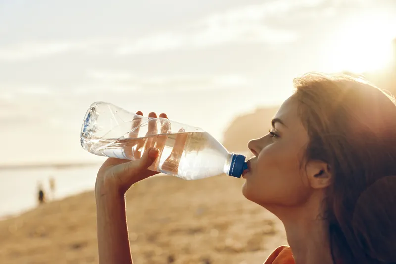 Woman drinking water from plastic bottle outdoors in desert, sunlight on blonde profile, dusty sand dunes behind, hydration and health focus, calm thirsty expression, fresh cool refreshment