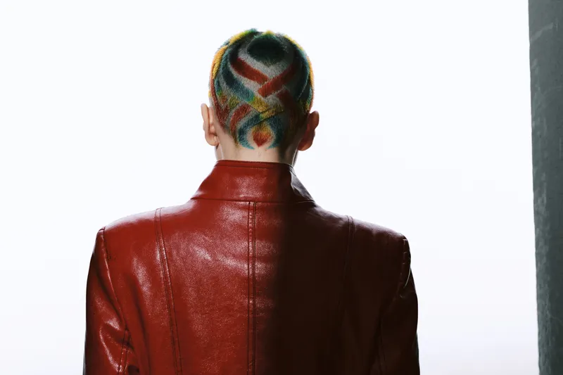 Woman with colorful geometric shaved hairstyle wearing a shiny red leather jacket standing against a bright white background, modern fashion look.