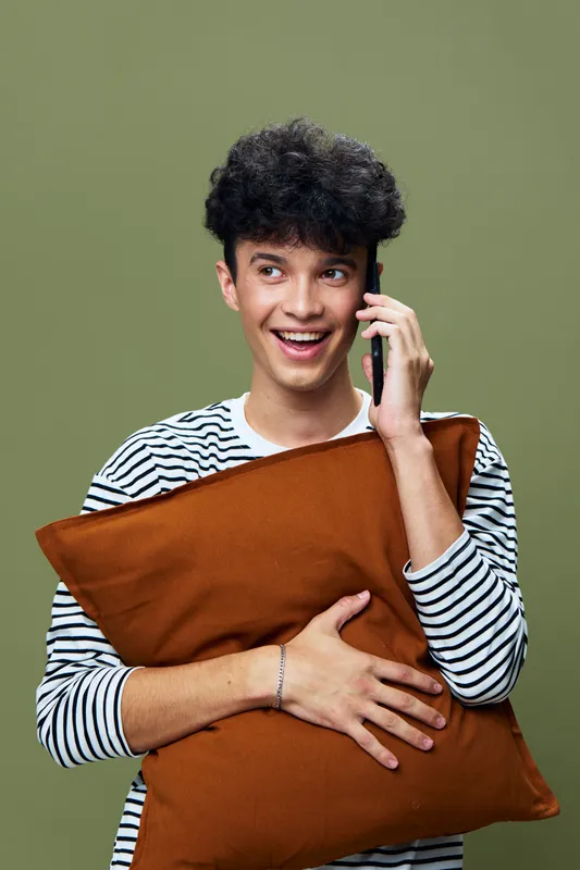 smiling man holding brown pillow and talking on smartphone, casual striped shirt, joyful expression, studio portrait with green background