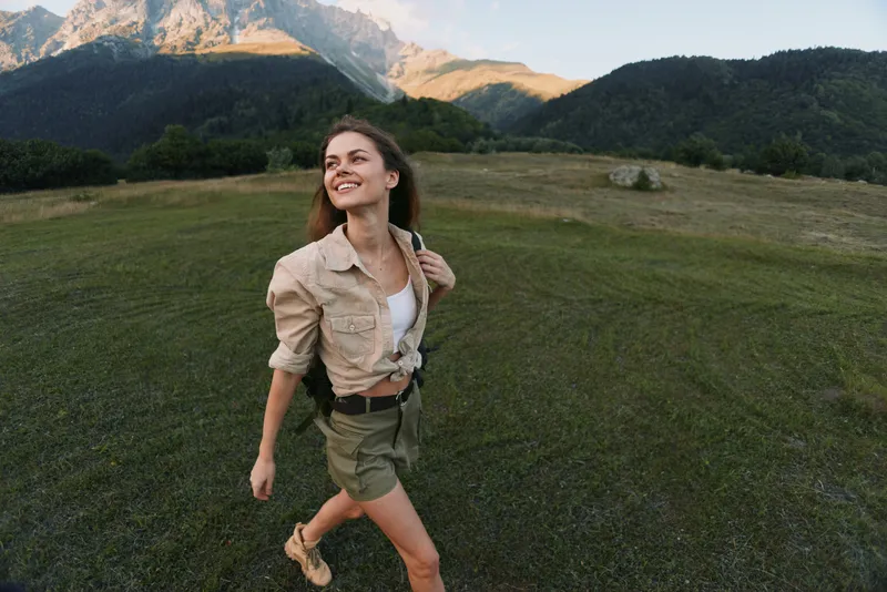 Young woman with long hair walks happily across a grassy field with mountains in the background during a clear day.