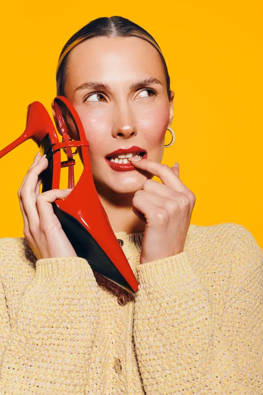 Young woman in yellow sweater holds red high heel shoe near face, biting finger, looking sideways on bright yellow background. Concept of fashion, style, and beauty.