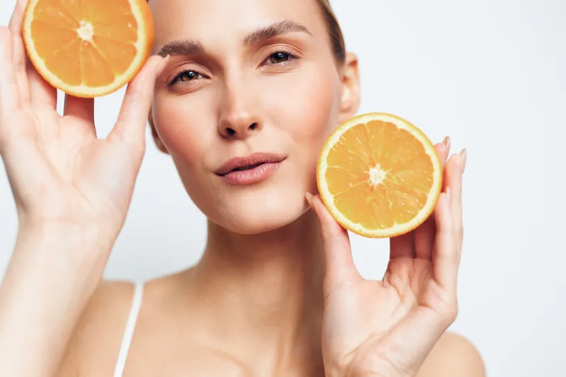 Young woman holds two halves of fresh orange near face shows natural skin beauty and healthy lifestyle on white background with positive expression and soft lighting close up portrait.
