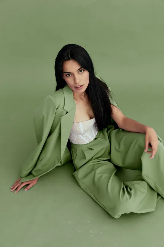 Young woman sitting on the floor wearing loose green pants and jacket over white top against matching green backdrop with serious expression, contemporary fashion style.