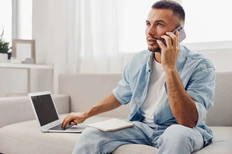 Engaged man on a phone call while working on a laptop, showcasing the modern blend of work-life balance in a cozy home environment with soft natural light.