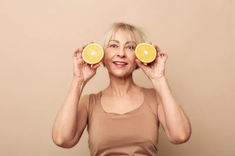 Happy mature woman holds two orange halves near her face smiling on beige background. Concept for healthy lifestyle, nutrition, natural vitamins, and joyful aging.