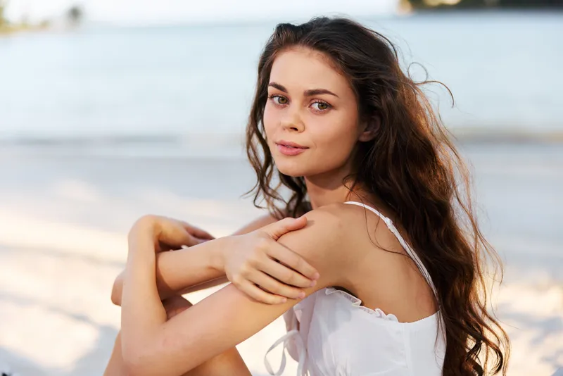 Portrait of a young woman on a sunlit beach, sitting with arms folded, wearing a white dress, soft gaze, and natural, relaxed expression.