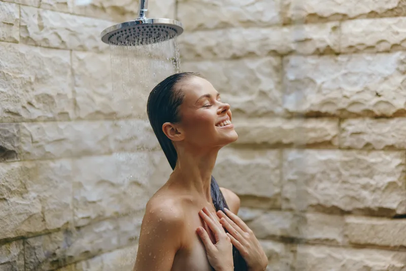 smiling woman enjoying outdoor shower with closed eyes under rain showerhead, natural stone wall background, relaxed and peaceful moment in sunlight