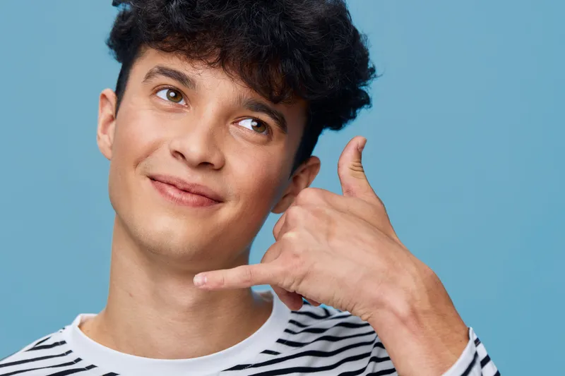 Young man in striped shirt making call me gesture with hand against a blue background, looking upward with a playful expression and curly hair.