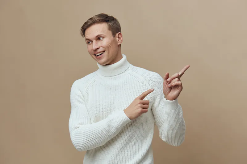 Smiling young man wearing white turtleneck sweater pointing to the side isolated on plain beige background studio portrait. People lifestyle concept