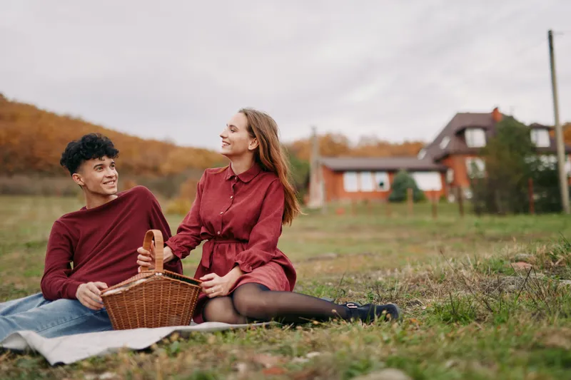 A young couple having a relaxed picnic on a grassy field by a rustic house, sharing genuine smiles and warm eye contact that reflect authenticity and credibility.