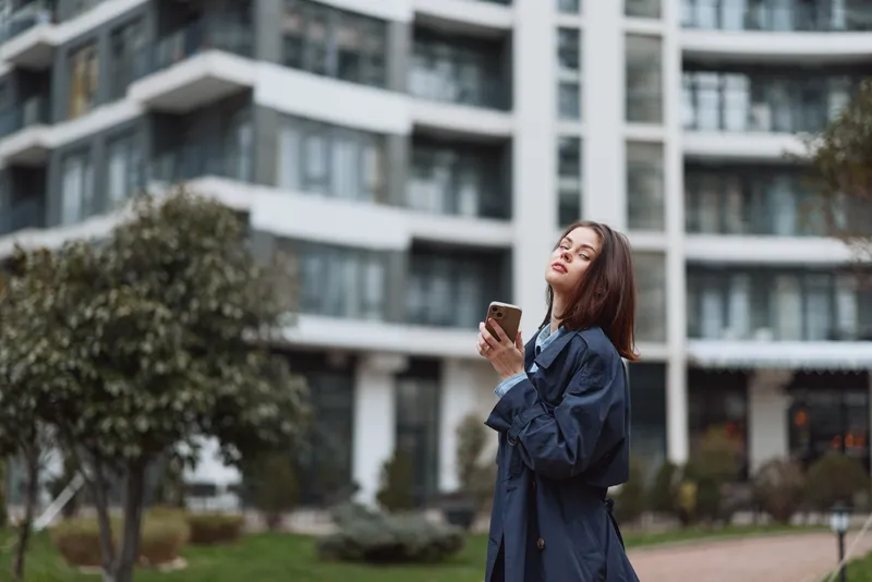 Young woman with dark hair wearing a denim jacket stands in a city courtyard, looking at her phone with a curious smile as modern buildings loom behind.