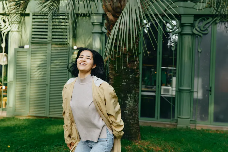 Smiling woman in a casual jacket outdoors among palms and greenery, enjoying a bright natural moment in a park setting, showcasing relaxed lifestyle for stock use