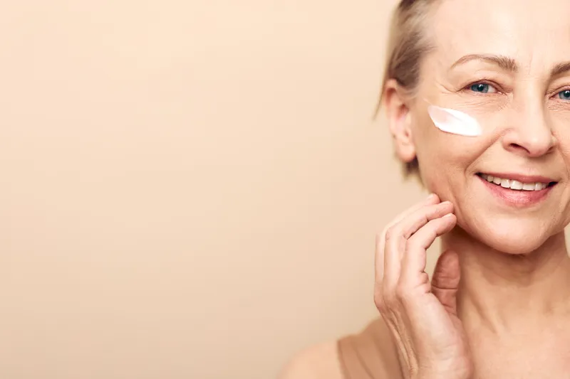 Smiling mature woman applies moisturizing cream on cheek, close-up on beige background. Concept of skincare, antiaging, beauty routine, healthy skin, wellness, and selfcare in natural light.