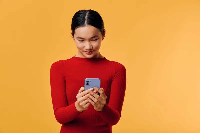 Young woman in red sweater standing against orange background using smartphone with focused expression, casual lifestyle, communication and technology concept.