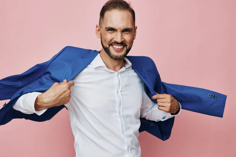 A professional man in a blue suit pulls his jacket aside with a confident smile against a soft pink isolated background, showcasing dynamic business energy and enthusiasm.