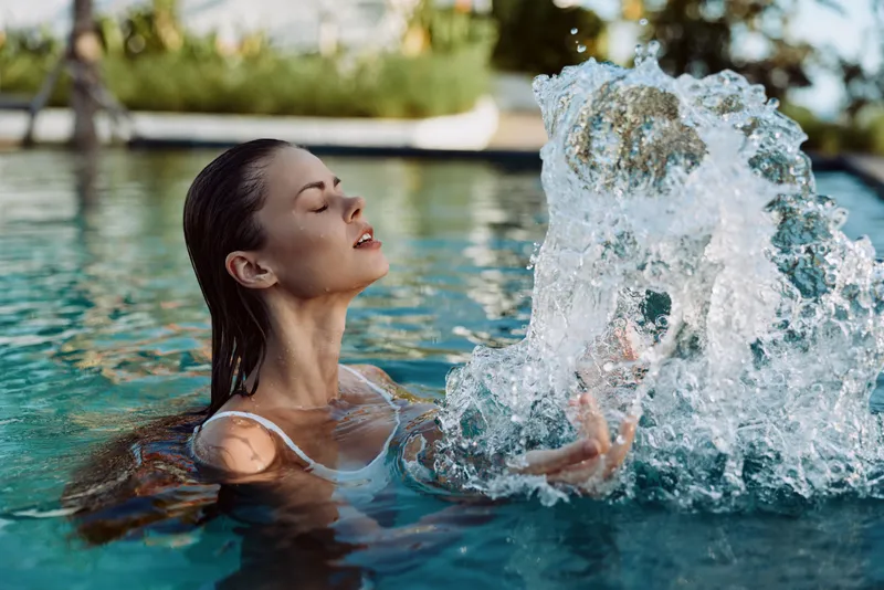 A woman enjoys a refreshing moment in a sunlit pool, her face relaxed as water splashes around her hands. Lifestyle scene highlighting leisure, fitness, and wellness by the water.