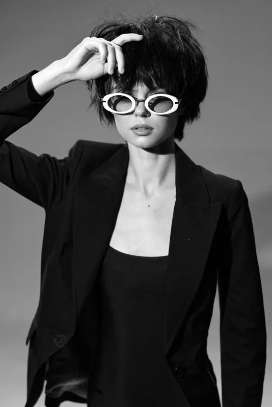Black and white portrait of a stylish woman wearing white sunglasses and a dark blazer with a confident expression and short hair.