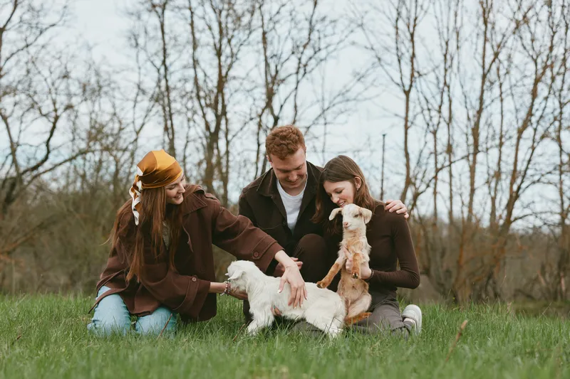 Three friends enjoy a sunny day in the park, kneeling to pet small dogs, sharing laughter and warm affection as casual jackets and green grass frame the simple outdoor moment.