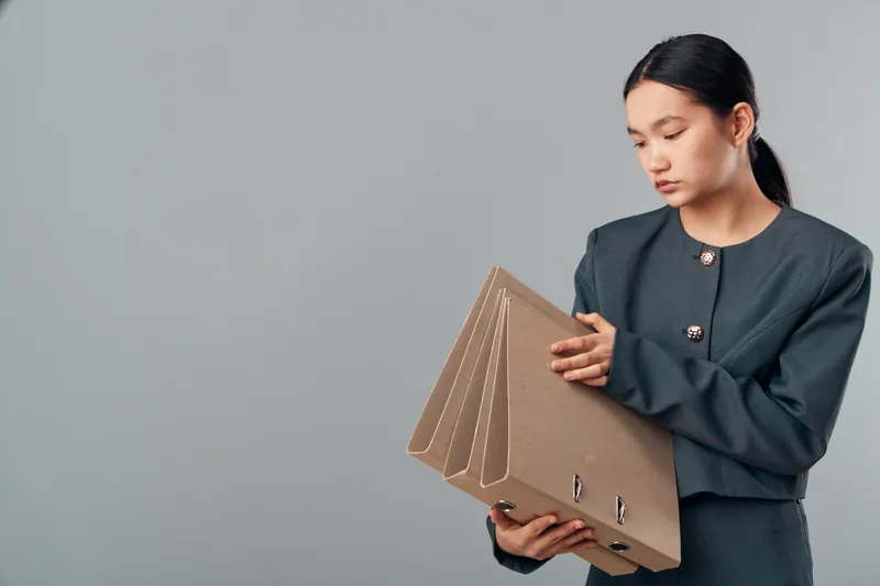 Woman in formal suit holding three brown cardboard file folders for organization, office work and document management with serious expression on gray background.