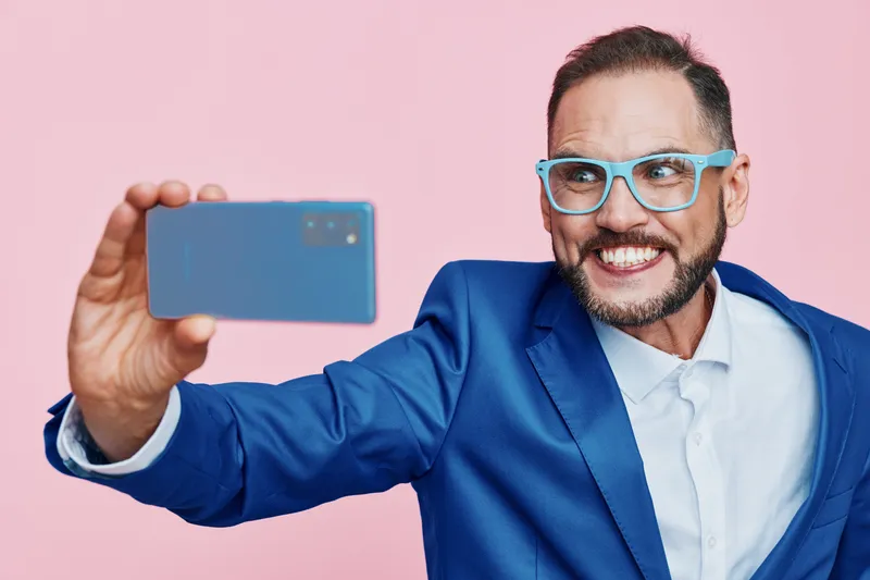A confident business professional man smiles while taking a selfie against a solid pink background, showcasing a modern, upbeat mood and clear, focused expression.